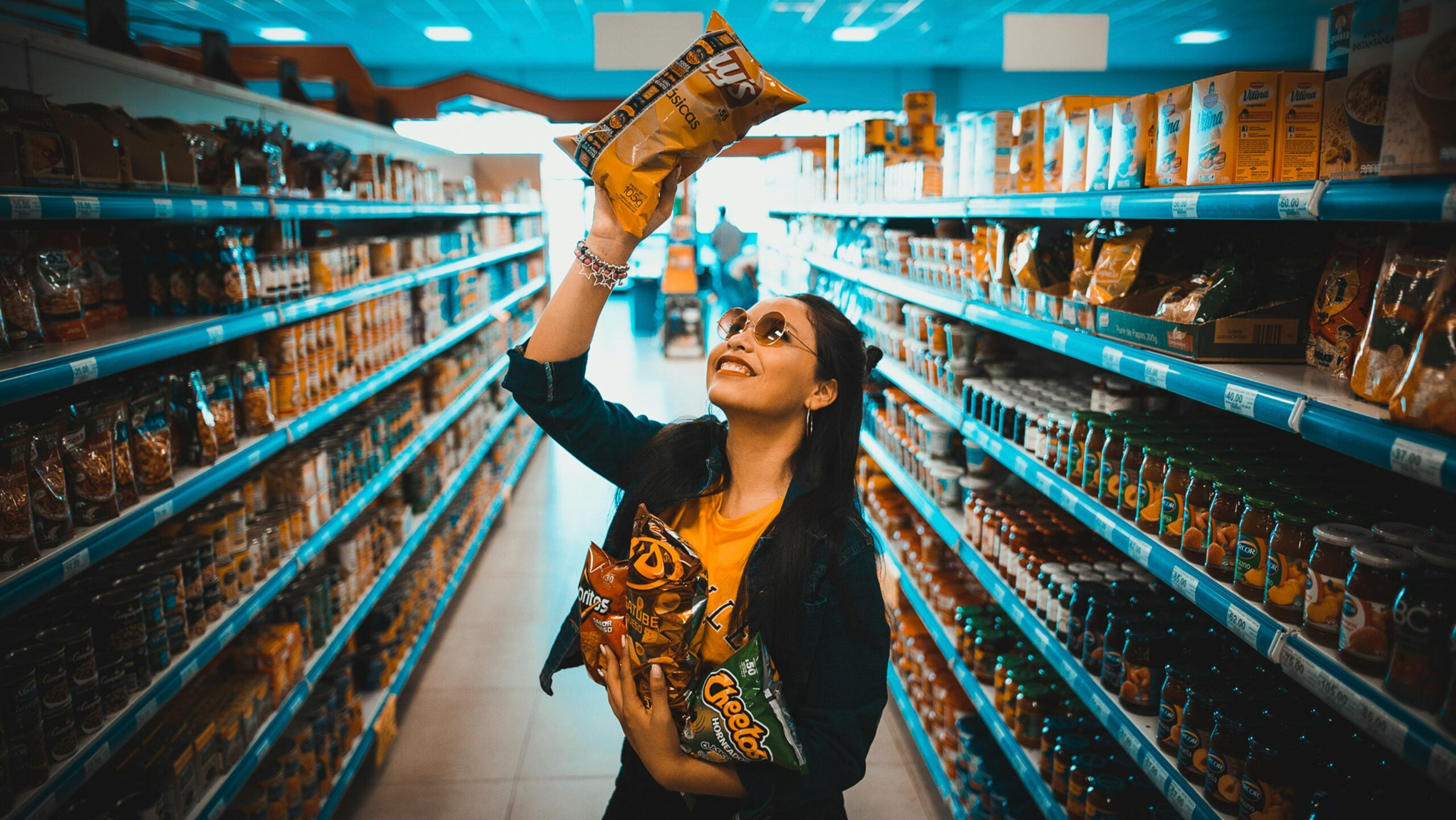 A woman gleefully holds bags of potato chips in a supermarket aisle, capturing a moment of joy while shopping.