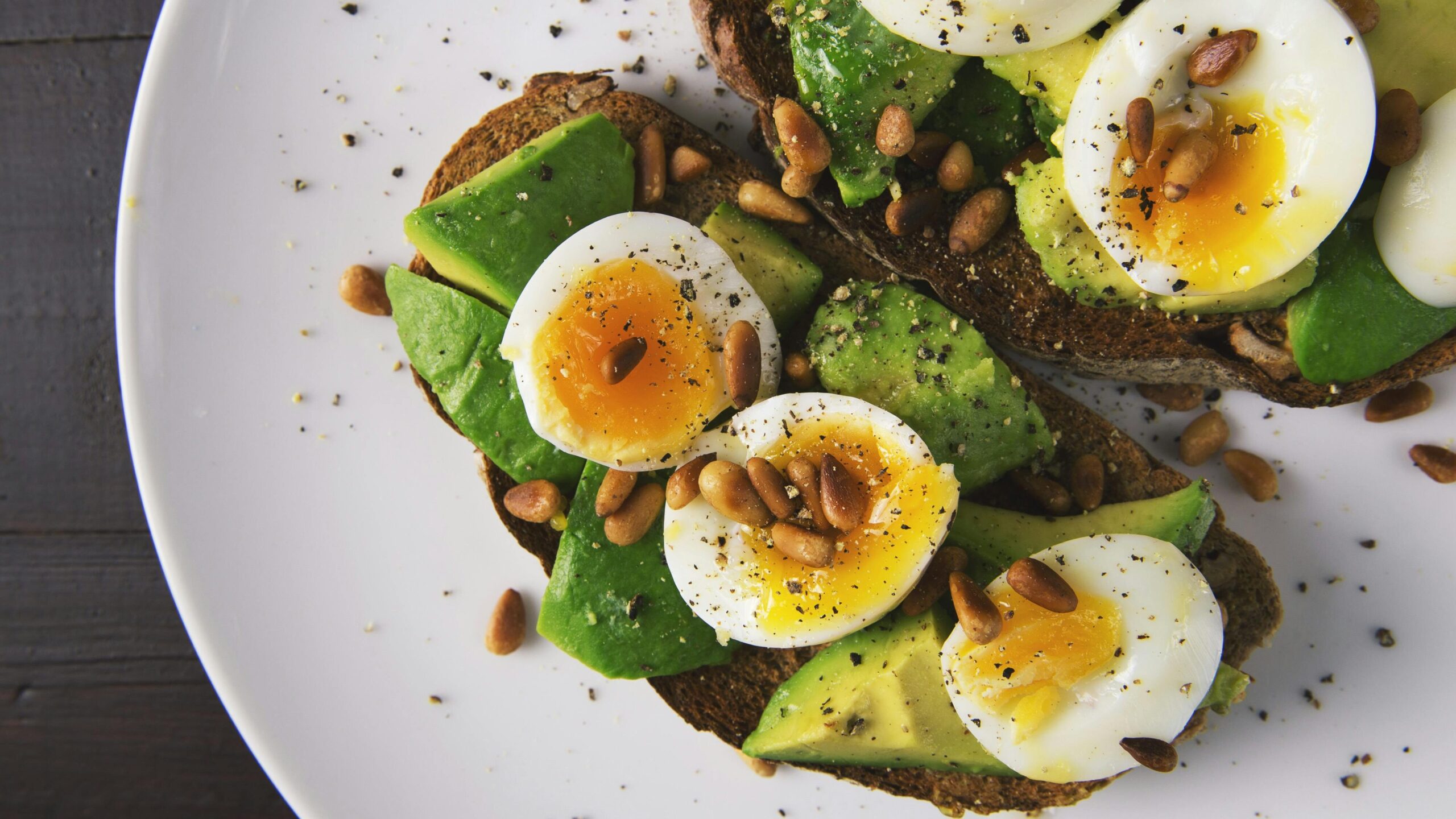Delicious avocado toast topped with soft-boiled eggs and pine nuts on a white plate.