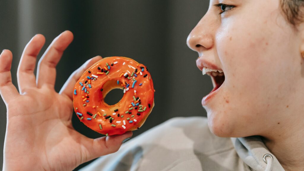 Crop glad female opening mouth to bite sweet glazed doughnut in light room